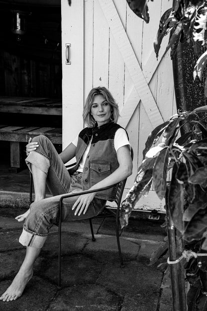 Woman in a broken twill denim vest and cuffed selvedge jeans seated on a metal chair by a white barn door.