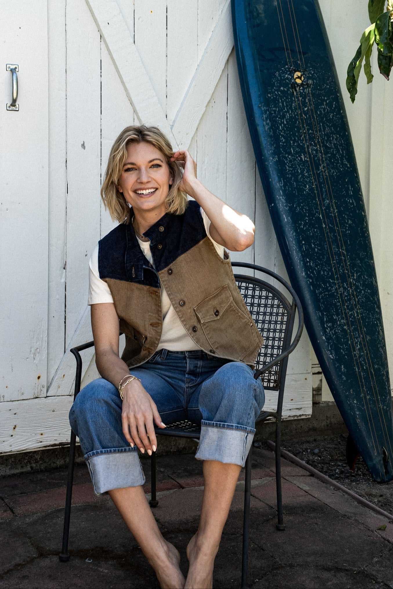 Woman seated on a metal chair wearing the wheat and indigo broken twill denim Jamie Vest with cuffed jeans, beside a.