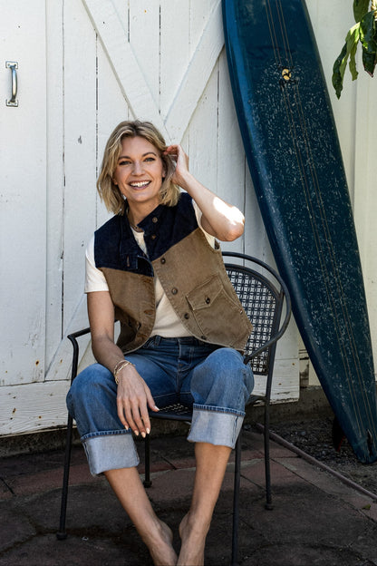 Woman seated on a metal chair wearing the wheat and indigo broken twill denim Jamie Vest with cuffed jeans, beside a.