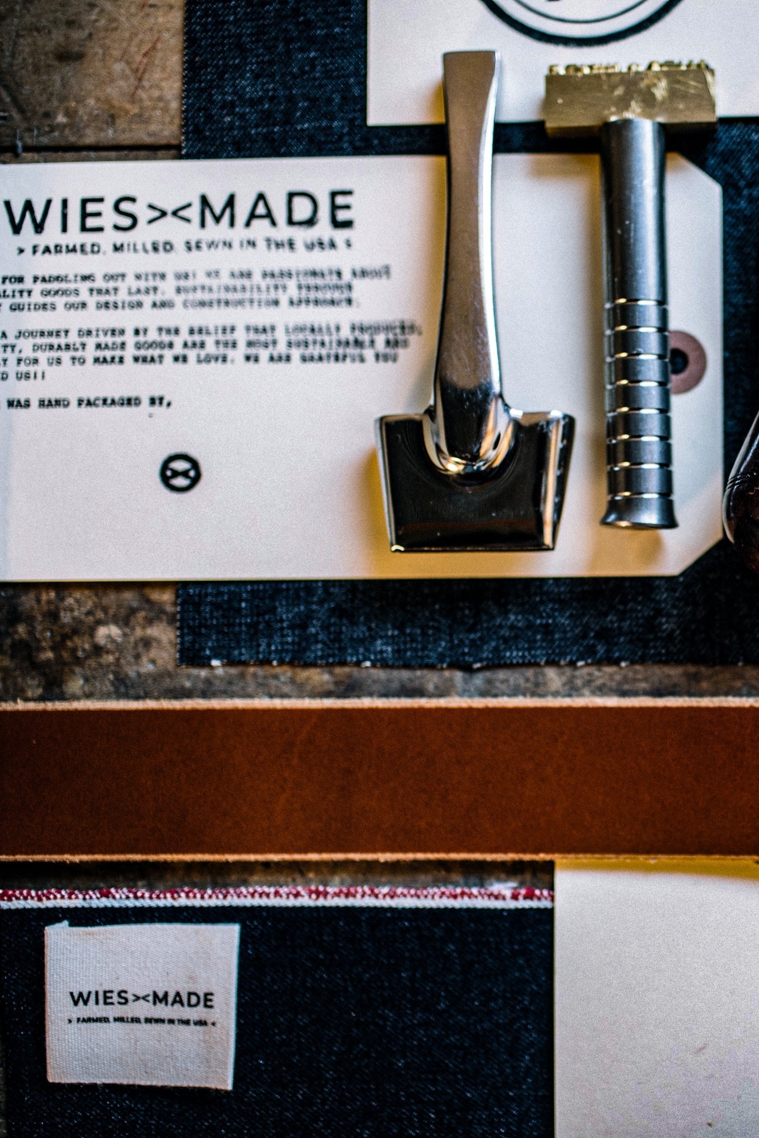 WiesMade flat-lay: tan Horween leather strip beside selvedge denim made in USA with brass stamping tools.