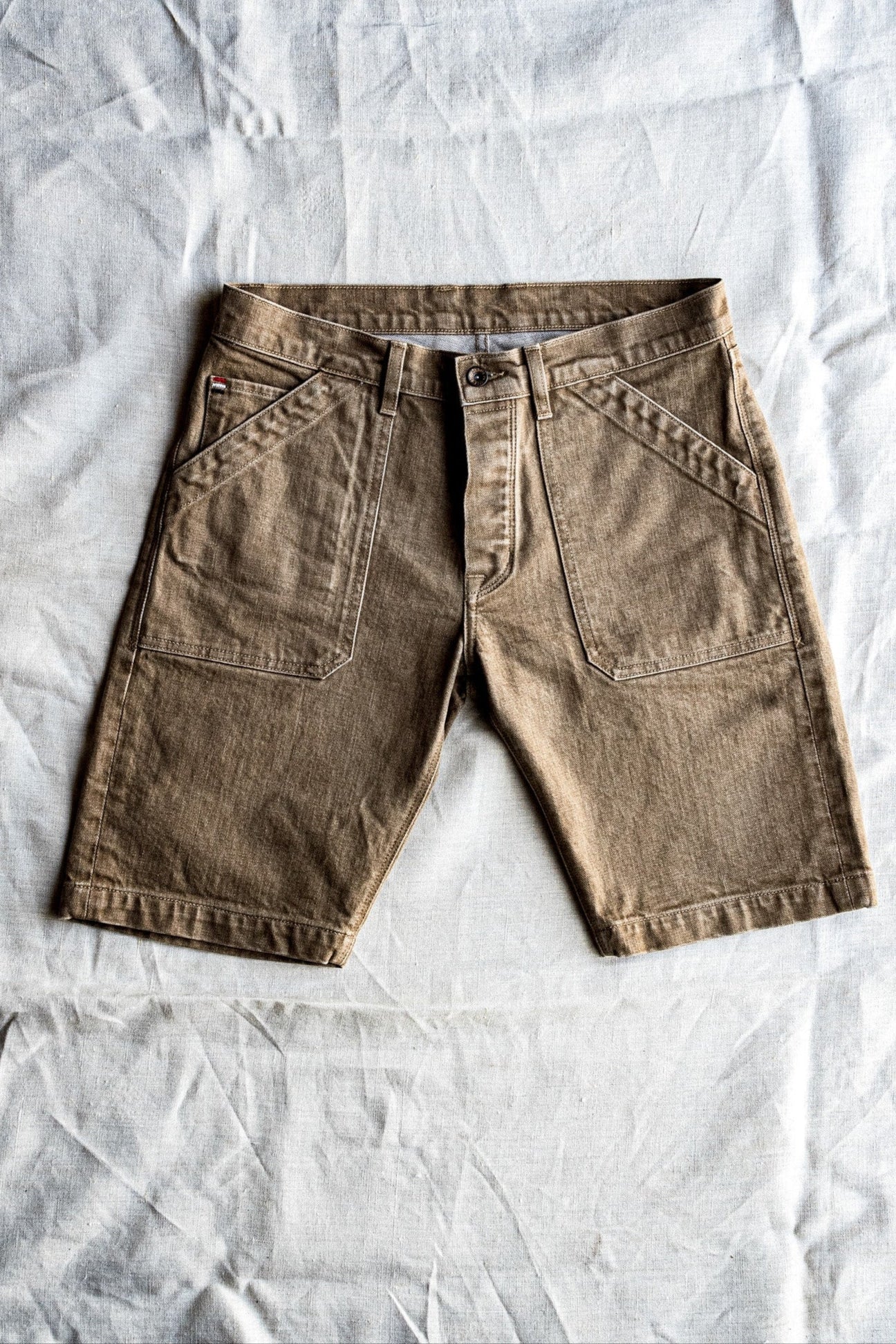 Wheat-colored broken twill denim military shorts laid flat on a linen backdrop, showing front patch pockets and button fly.