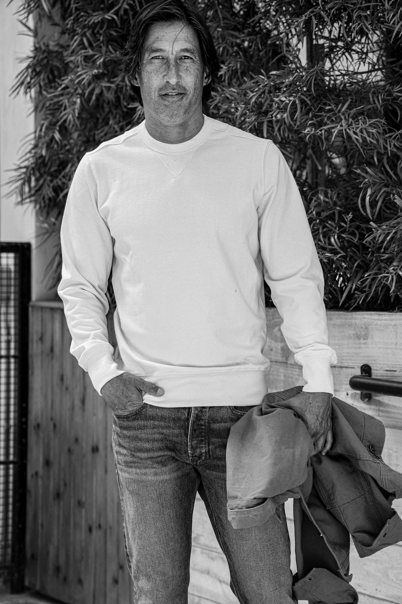 Man wearing a natural white Stinson crewneck pullover with jeans, holding a jacket, outdoors by foliage.