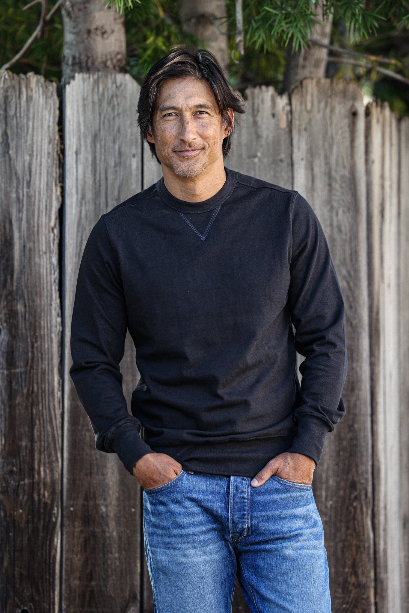 Man wearing the basalt black Stinson rugby jersey pullover with blue jeans, standing by a weathered wood fence.