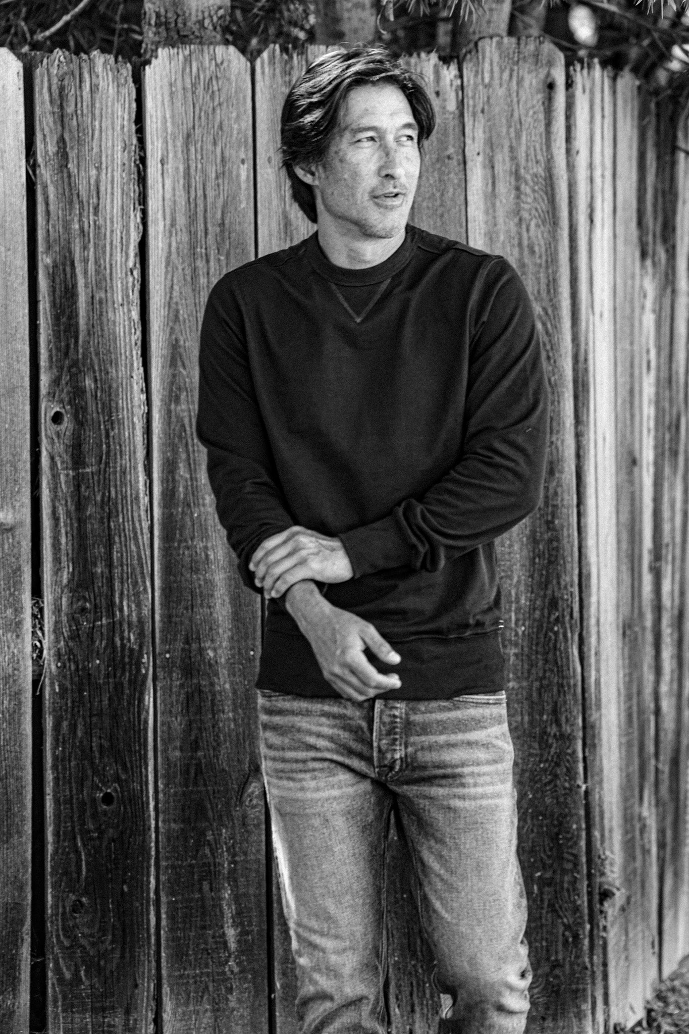 Man in a dark Stinson rugby jersey pullover and jeans leaning against a weathered wood fence.
