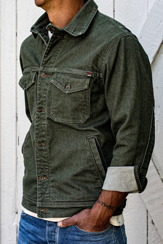 Side view of olive broken twill denim jacket with chest pocket, brass buttons, and cuffed sleeve against white wood wall.