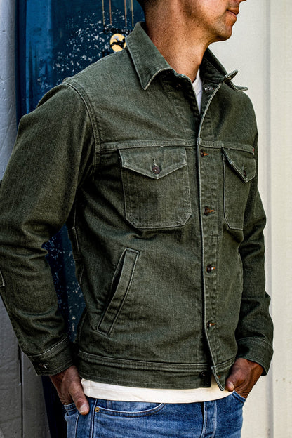 Side view of a man wearing the olive broken twill denim jacket with chest pockets and brass buttons.