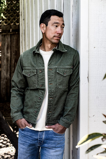Man in olive broken twill denim jacket with chest pockets and brass buttons, leaning against a white wall.