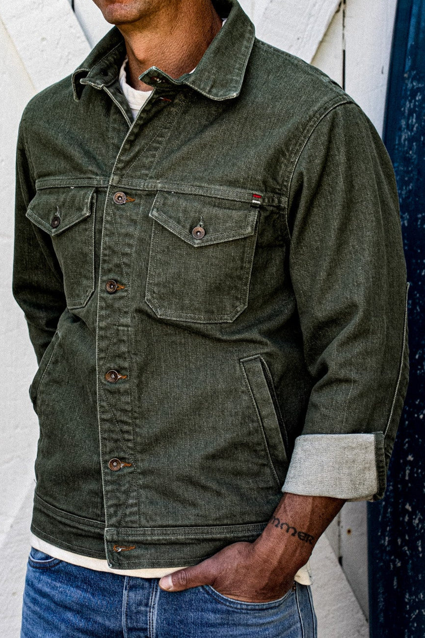Olive broken twill denim jacket worn buttoned over a cream tee with one cuff rolled to show the selvedge edge.