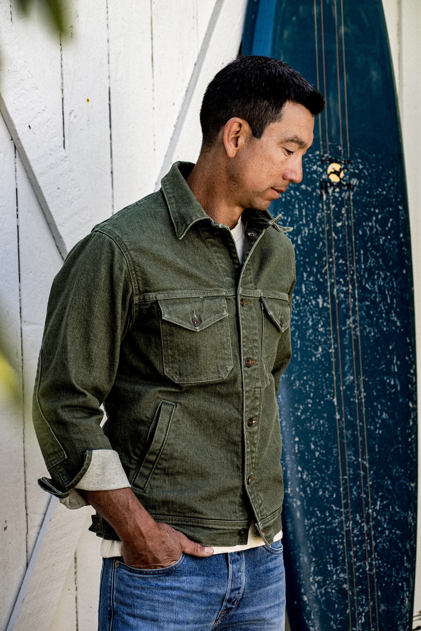Man wearing olive broken twill denim jacket with flap chest pockets and rolled cuffs, beside a surfboard.