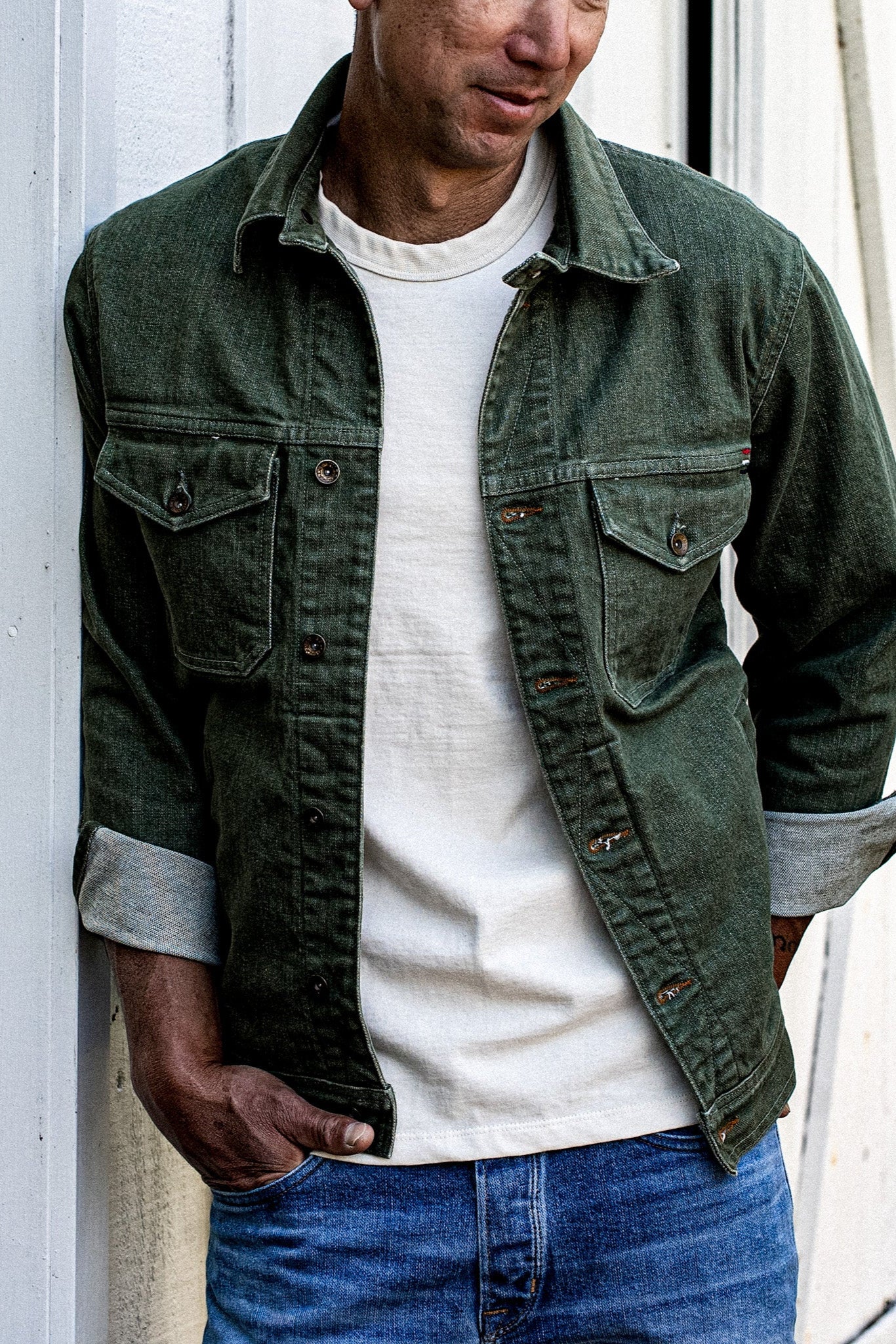 Man in an olive broken twill denim jacket with cuffed sleeves over a white tee and blue jeans.