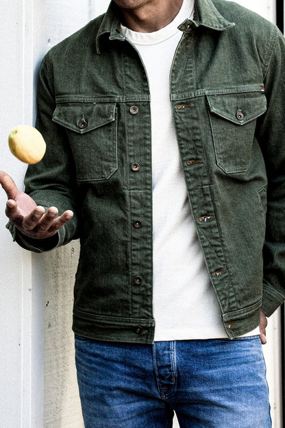Olive broken twill denim jacket worn open over a white tee with jeans, tossing a lemon.