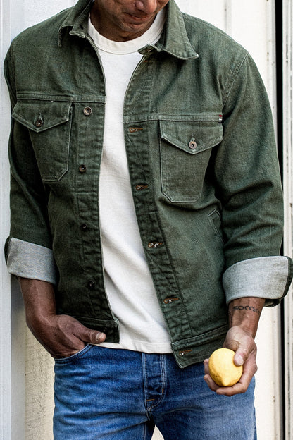 Model wearing the olive broken twill denim jacket open over a white tee with blue jeans, cuffs rolled.