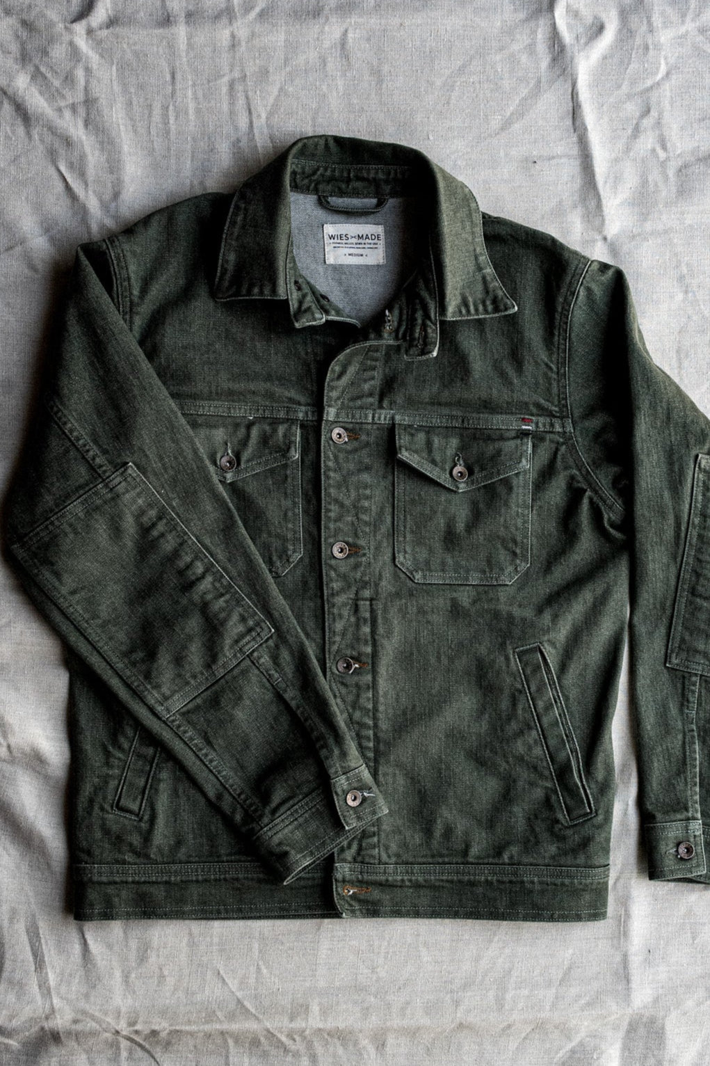 Olive broken twill denim jacket laid flat, showing collar, chest pockets with brass buttons, and welt hand pockets.
