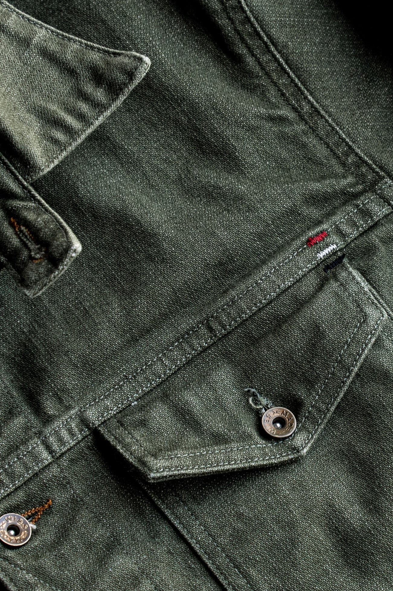 Close-up of olive broken twill denim jacket showing chest pocket, flat felled seams, and brass buttons.