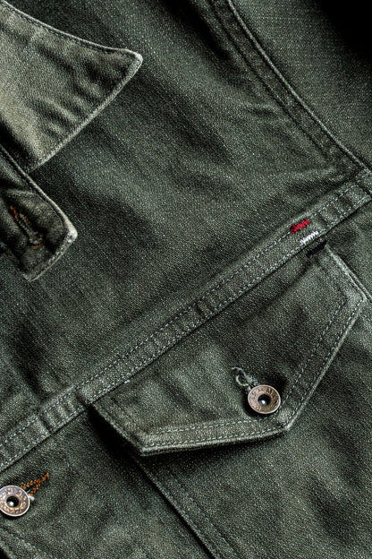 Close-up of olive broken twill denim jacket showing chest pocket, flat felled seams, and brass buttons.