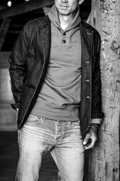 Man in Bodega shawl collar French terry pullover layered under a denim blazer, leaning on a wooden post.