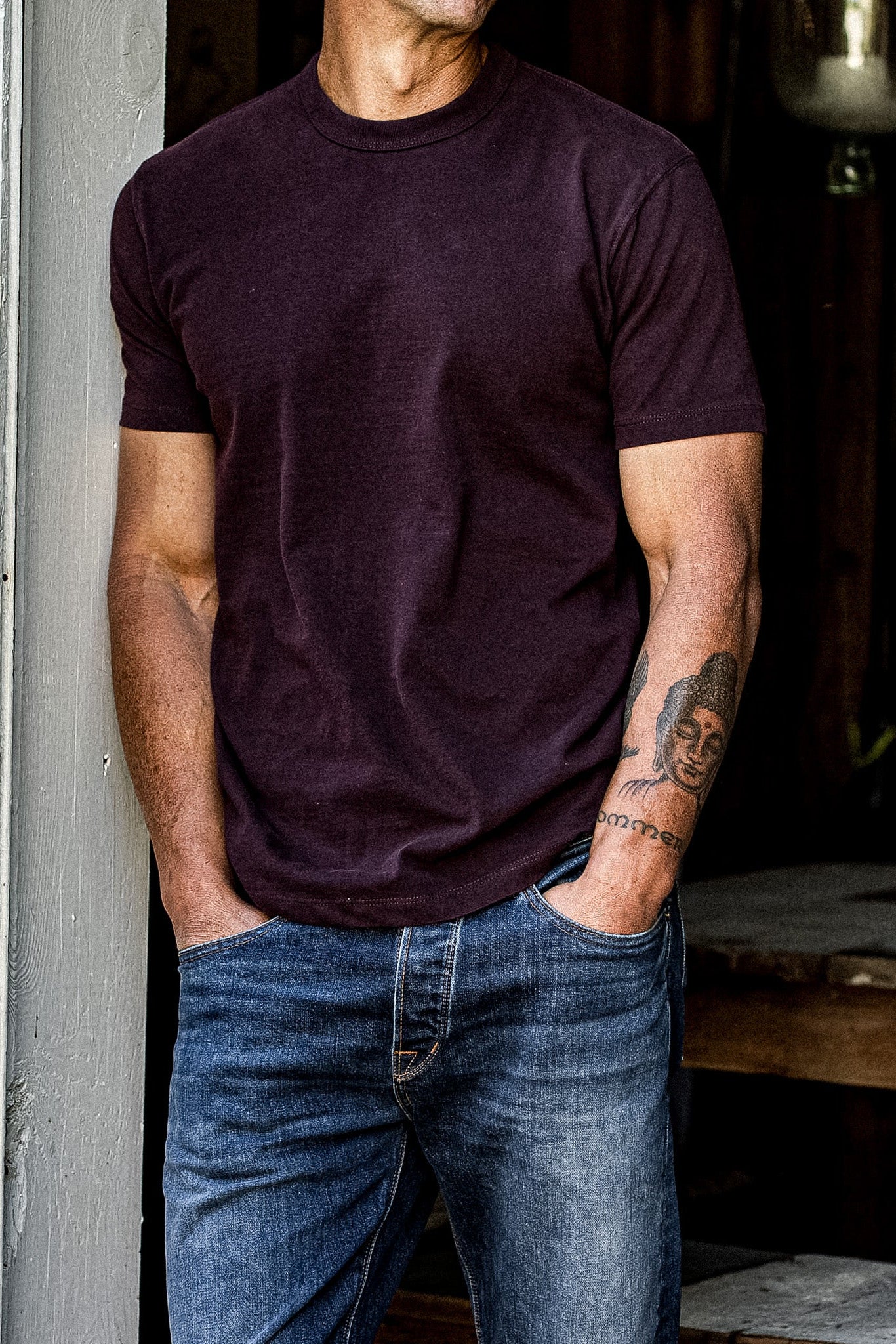 Oxblood heavyweight cotton tee worn with faded blue jeans, leaning in a doorway.