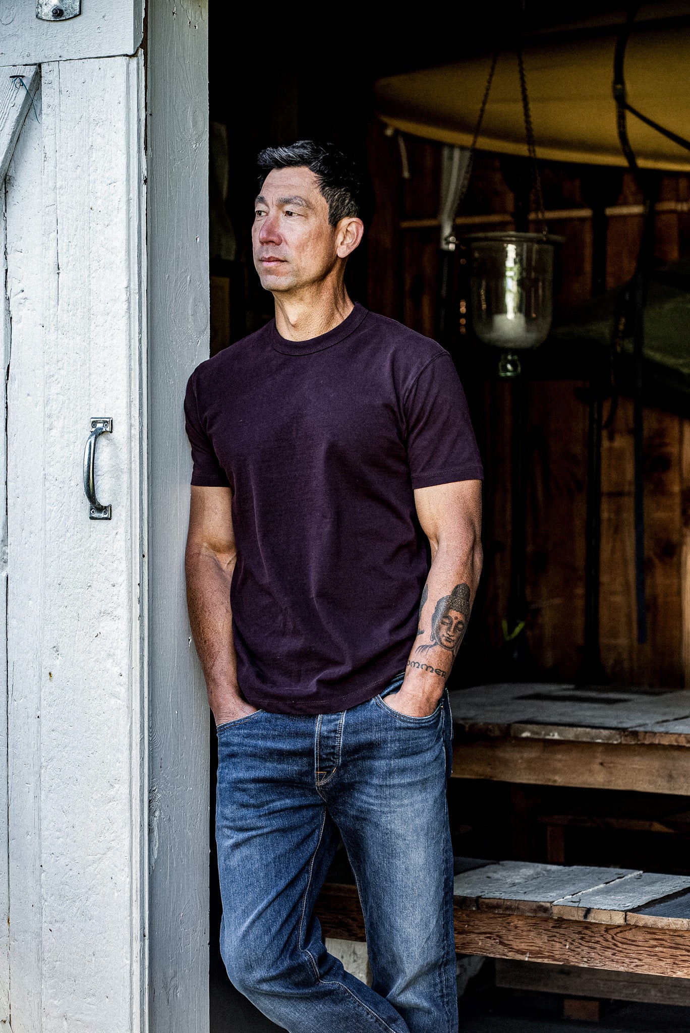 Man in oxblood heavyweight cotton Bolinas tee and jeans leaning against a weathered doorway.