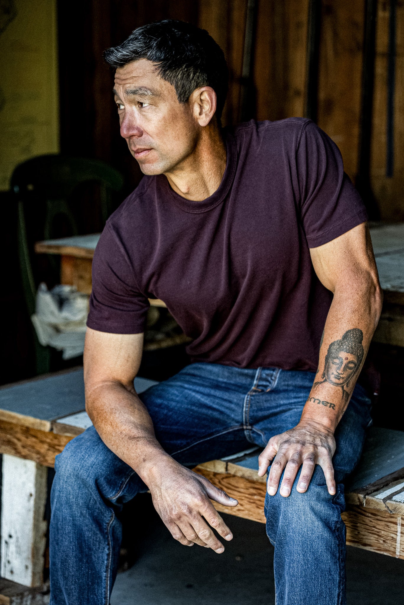 Model wears the oxblood Bolinas heavyweight cotton tee with jeans, seated in a rustic workshop.
