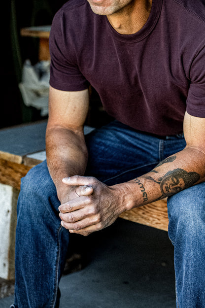 Seated man in an oxblood heavyweight cotton tee, worn with dark denim jeans.
