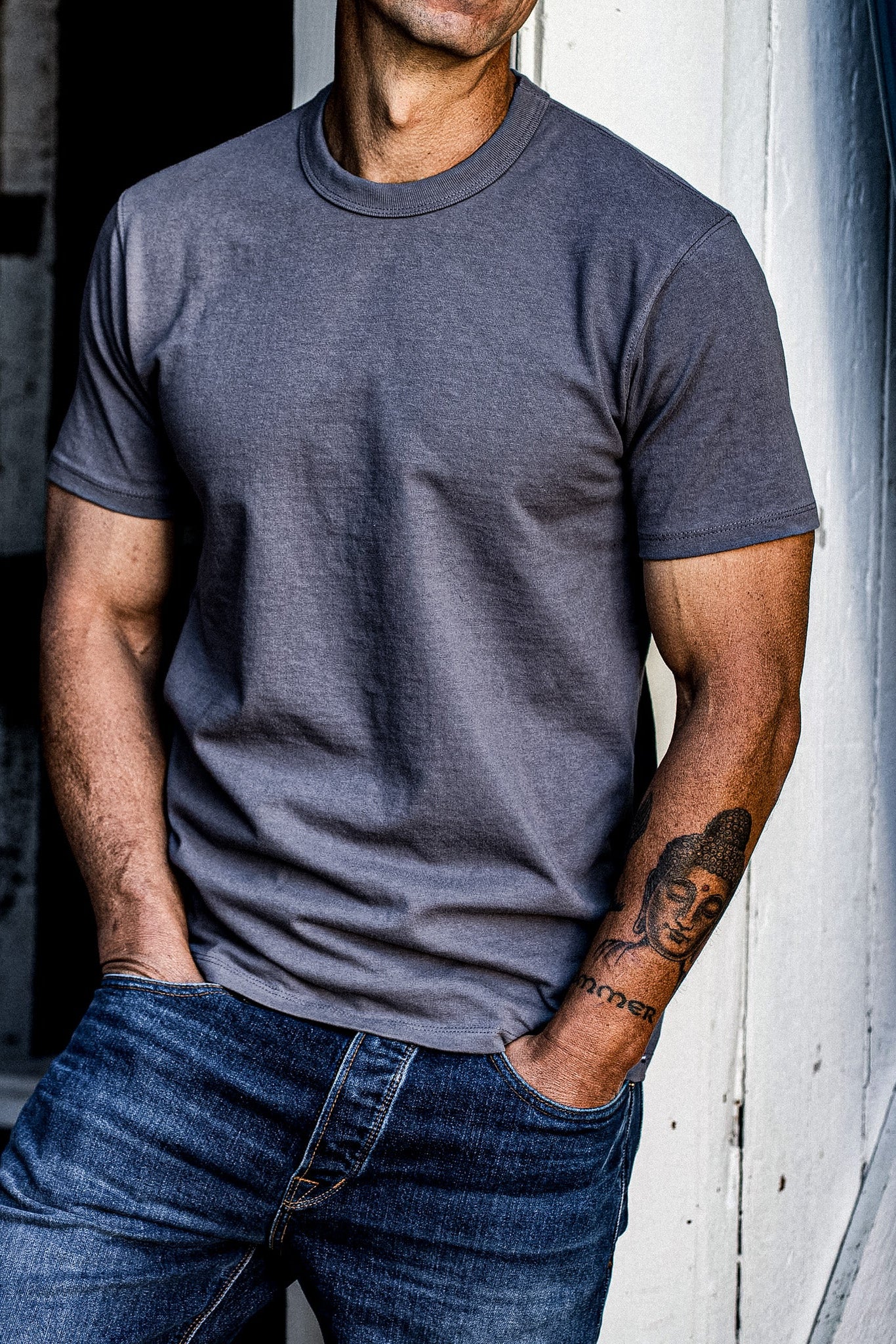 Person wearing pebble grey heavyweight cotton Bolinas tee with jeans, leaning on white wall.