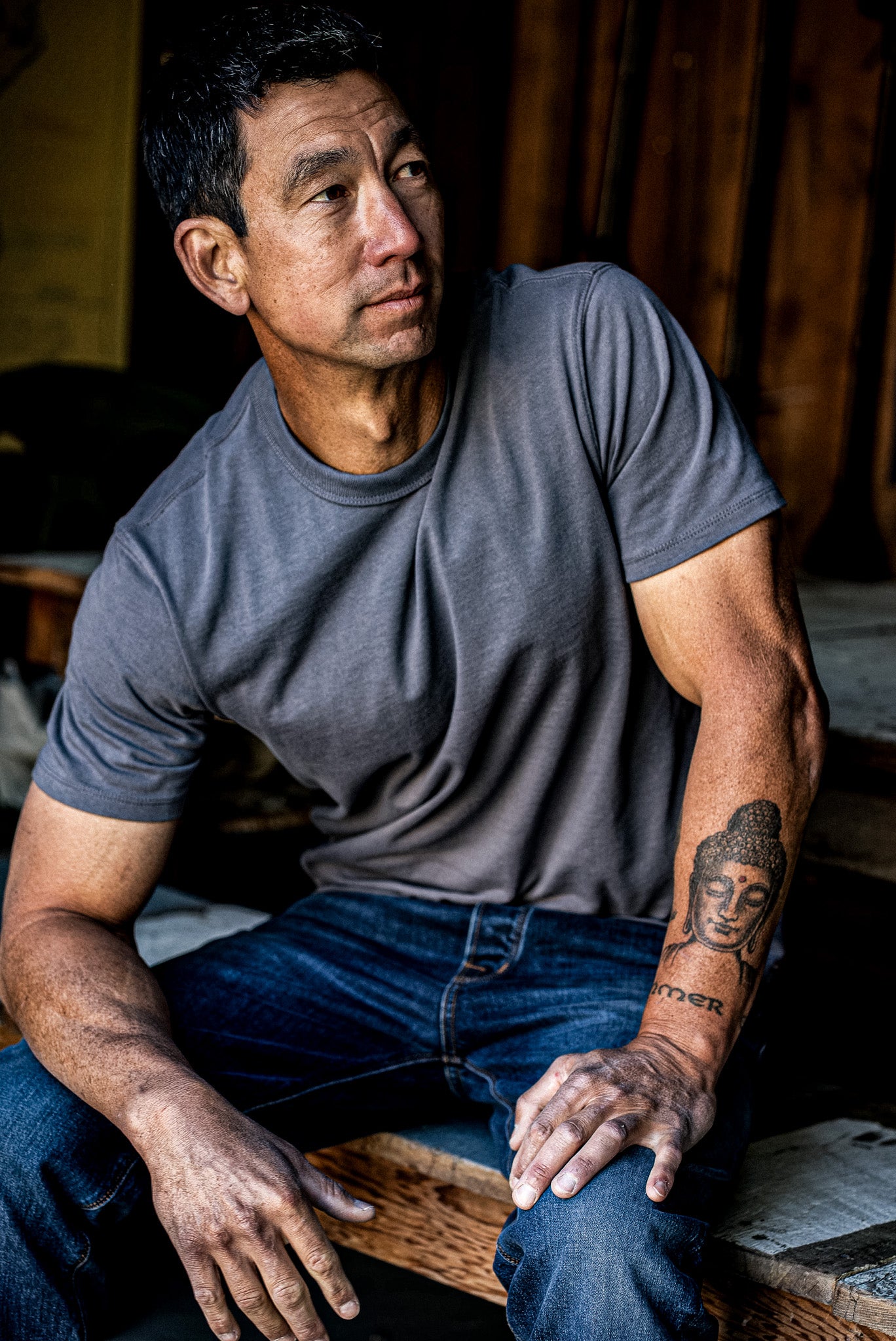 Man wearing pebble gray t-shirt and jeans, seated with visible arm tattoo