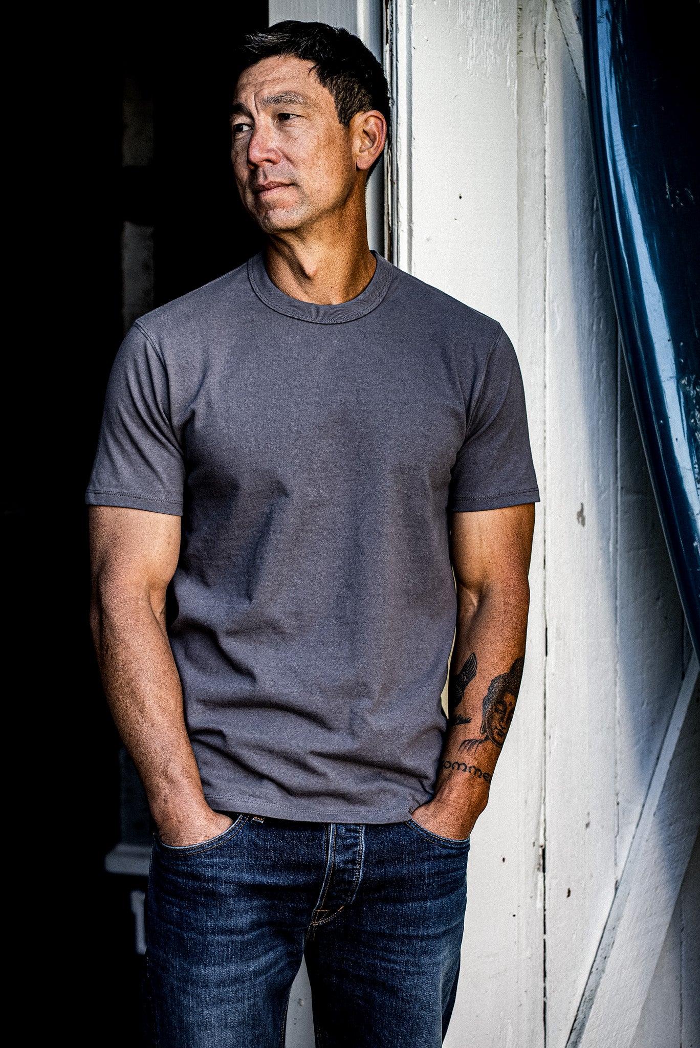 Man in pebble grey Bolinas heavyweight cotton tee and jeans by a white doorway.