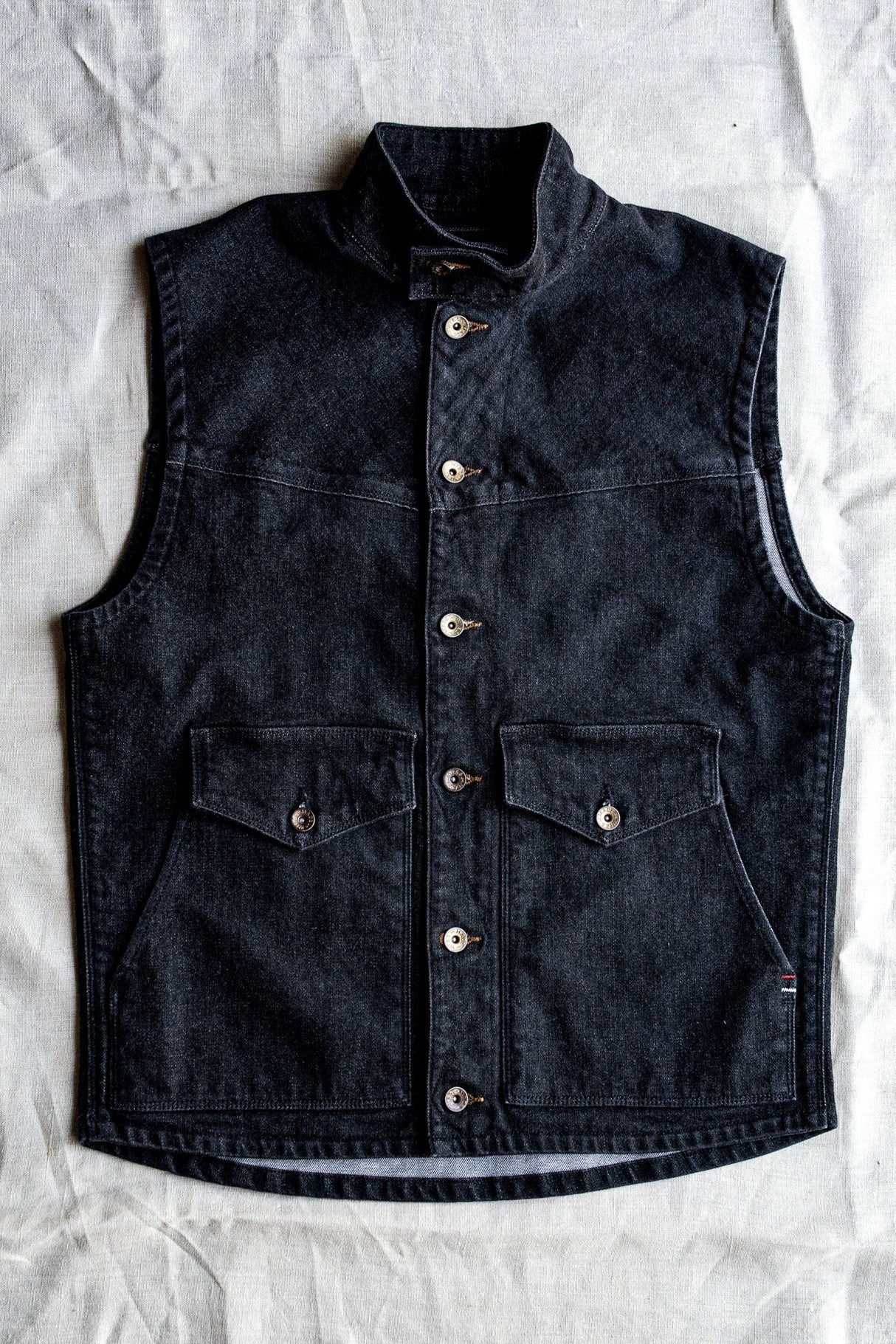 Black broken twill denim vest with stand collar, brass snap buttons, and flap chest pockets, laid flat on linen.