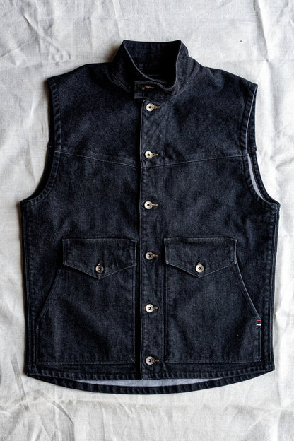 Black broken twill denim vest with stand collar, brass snap buttons, and flap chest pockets, laid flat on linen.