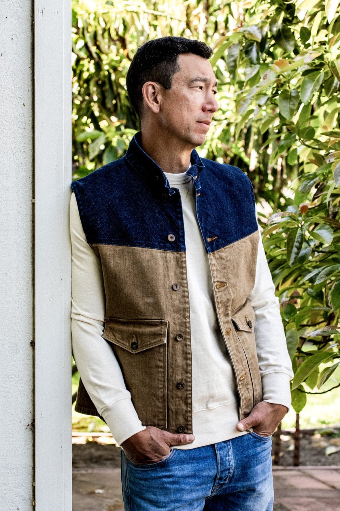Man wearing wheat and blue broken twill denim vest over cream sweatshirt, leaning against a post outdoors.