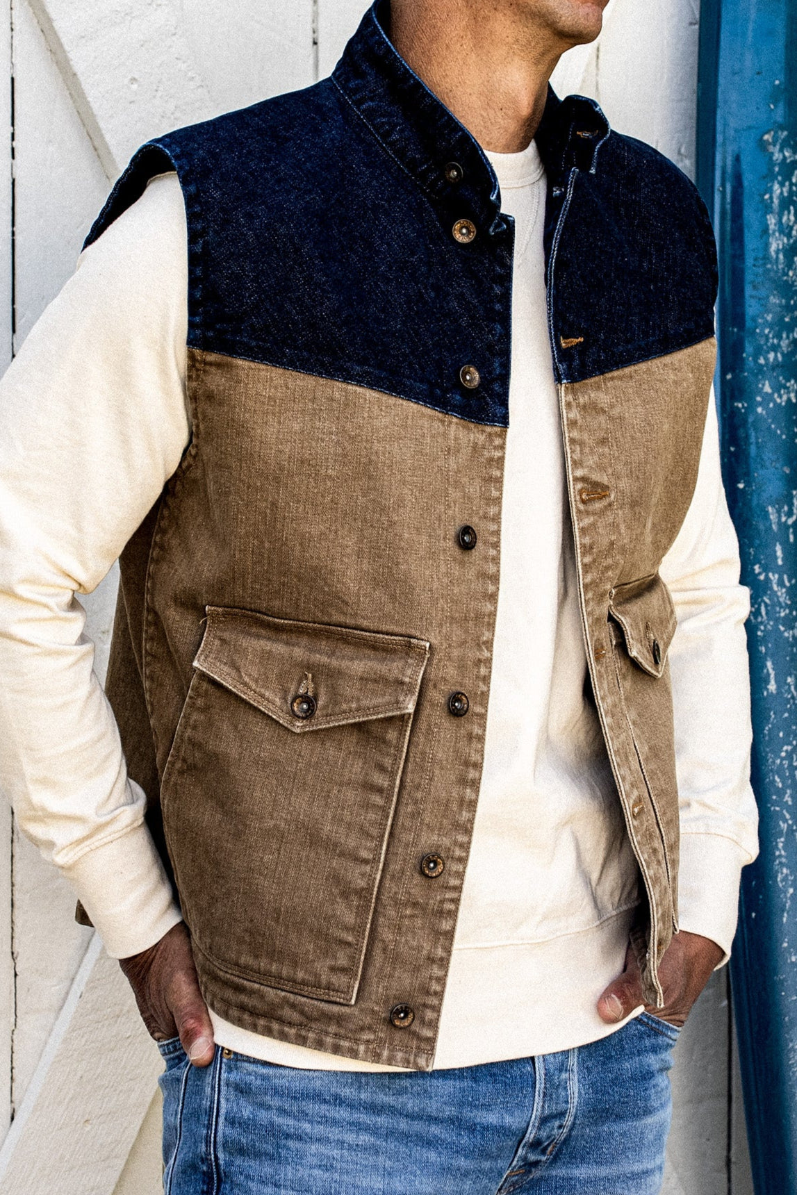 Two-tone broken twill denim vest with wheat body and indigo yoke, worn over a cream sweatshirt.