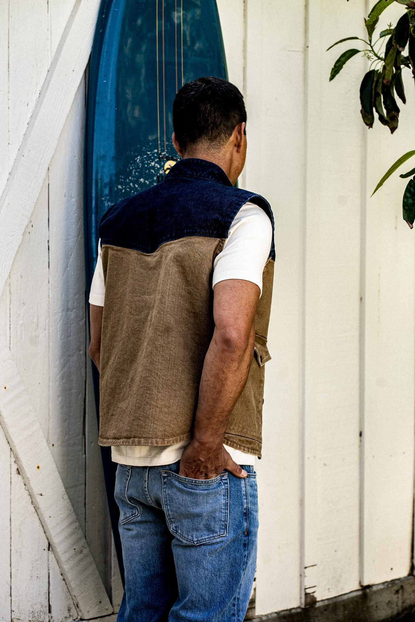 Back view of the James Vest in wheat broken twill denim with indigo yoke, worn with jeans beside a surfboard.