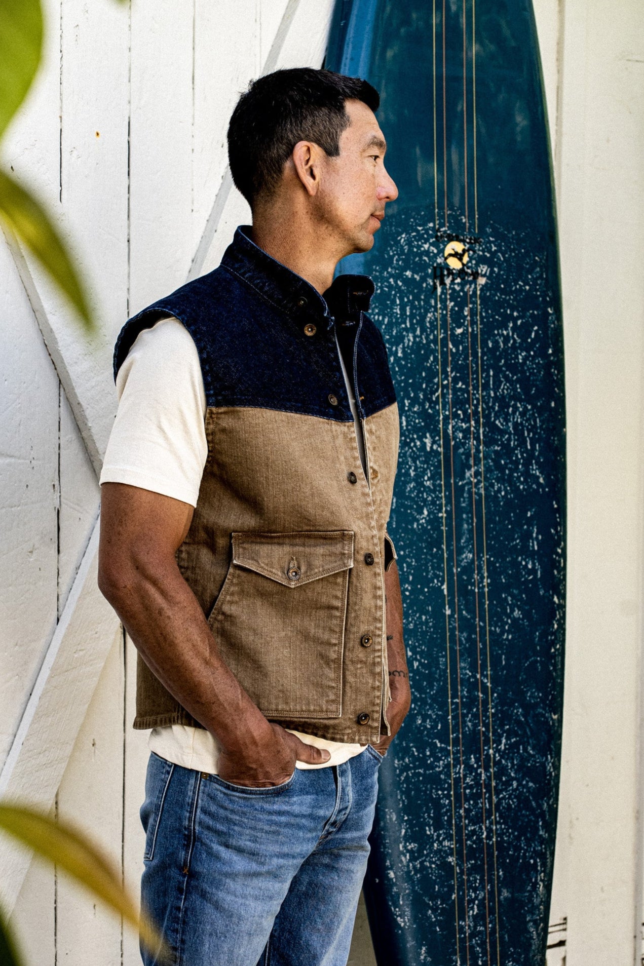Man wearing the James Vest in wheat and indigo broken twill denim beside a surfboard.