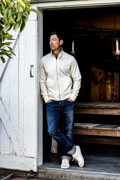 Man in a natural cotton moto zip jacket leaning in a rustic barn doorway.