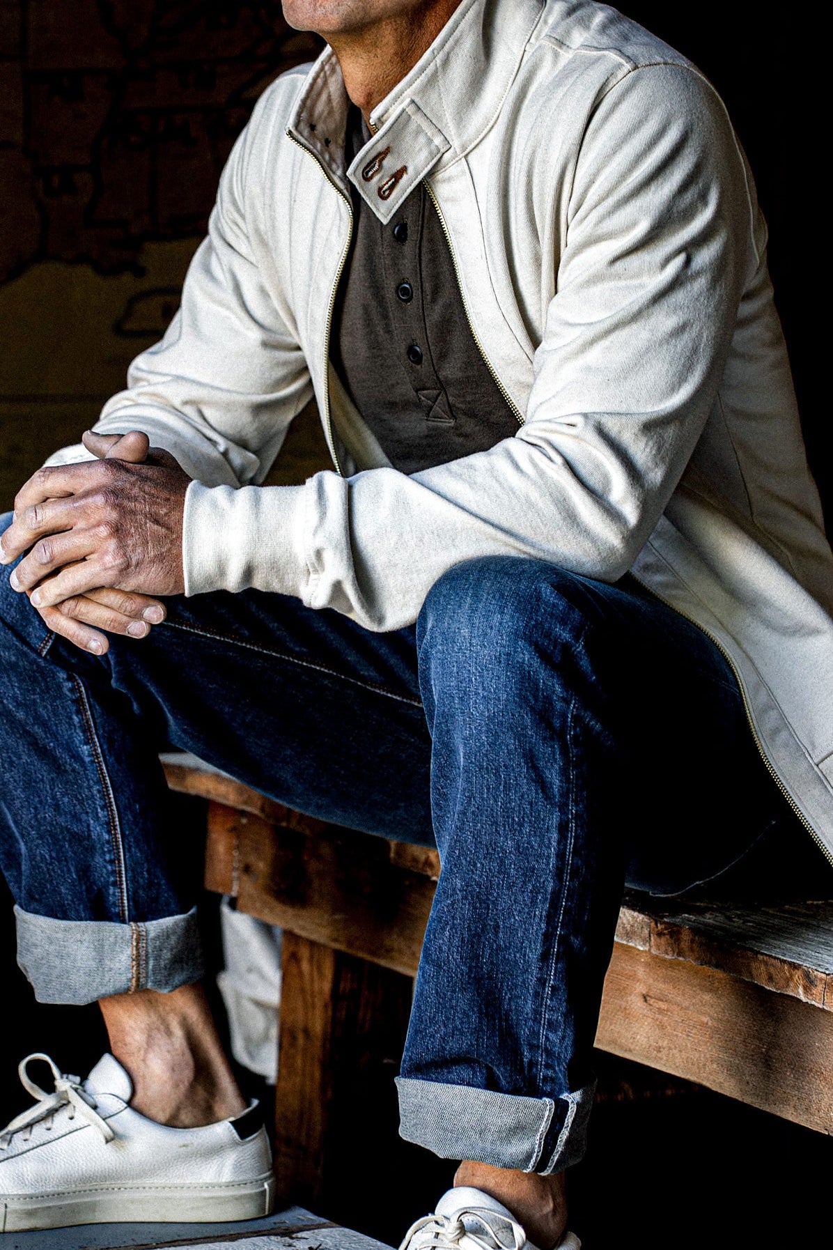 Seated man wearing a natural cotton moto zip jacket over cuffed jeans and white sneakers.