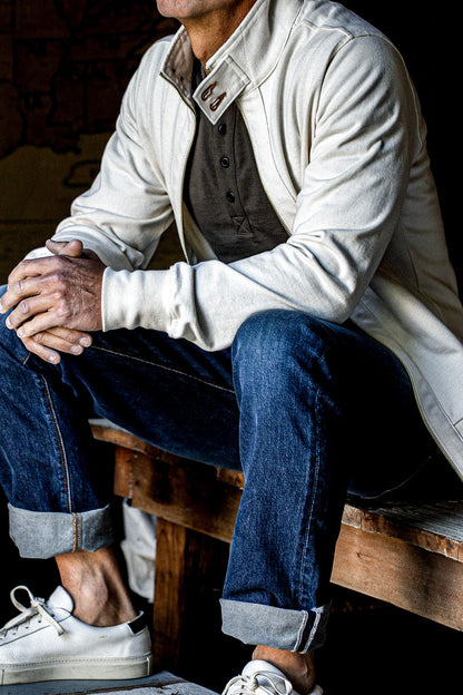 Seated man wearing a natural cotton moto zip jacket over cuffed jeans and white sneakers.