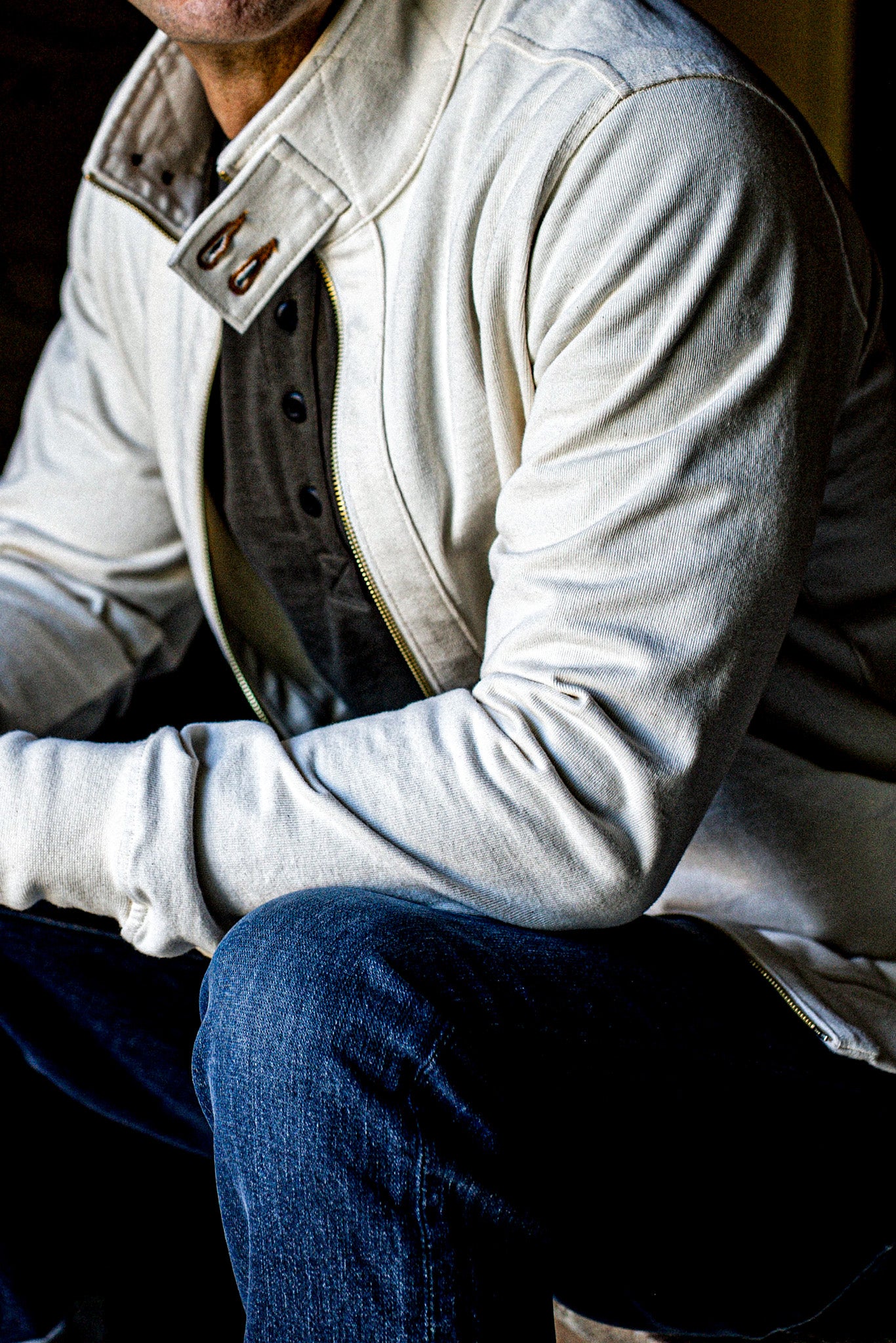 Man seated in a natural cotton moto zip jacket with brass zipper and blue jeans.