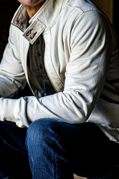 Man seated in a natural cotton moto zip jacket with brass zipper and blue jeans.