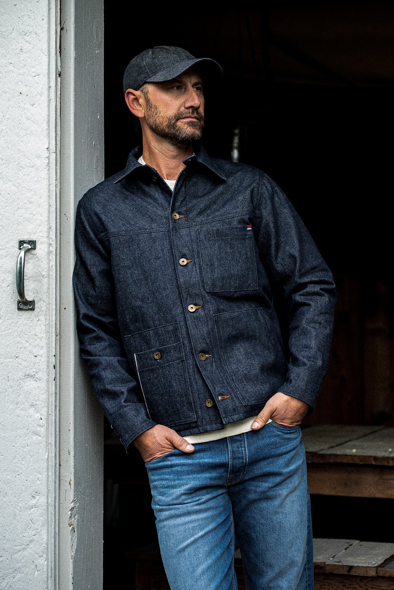 Man in the WiesMade Nico indigo denim jacket and ball cap leaning in a doorway, upper body view.