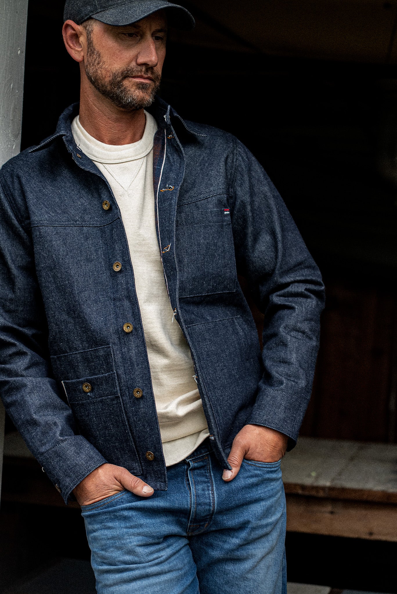 Man wearing the WiesMade Nico indigo denim jacket open with a ball cap and cream sweatshirt, upper body shot.