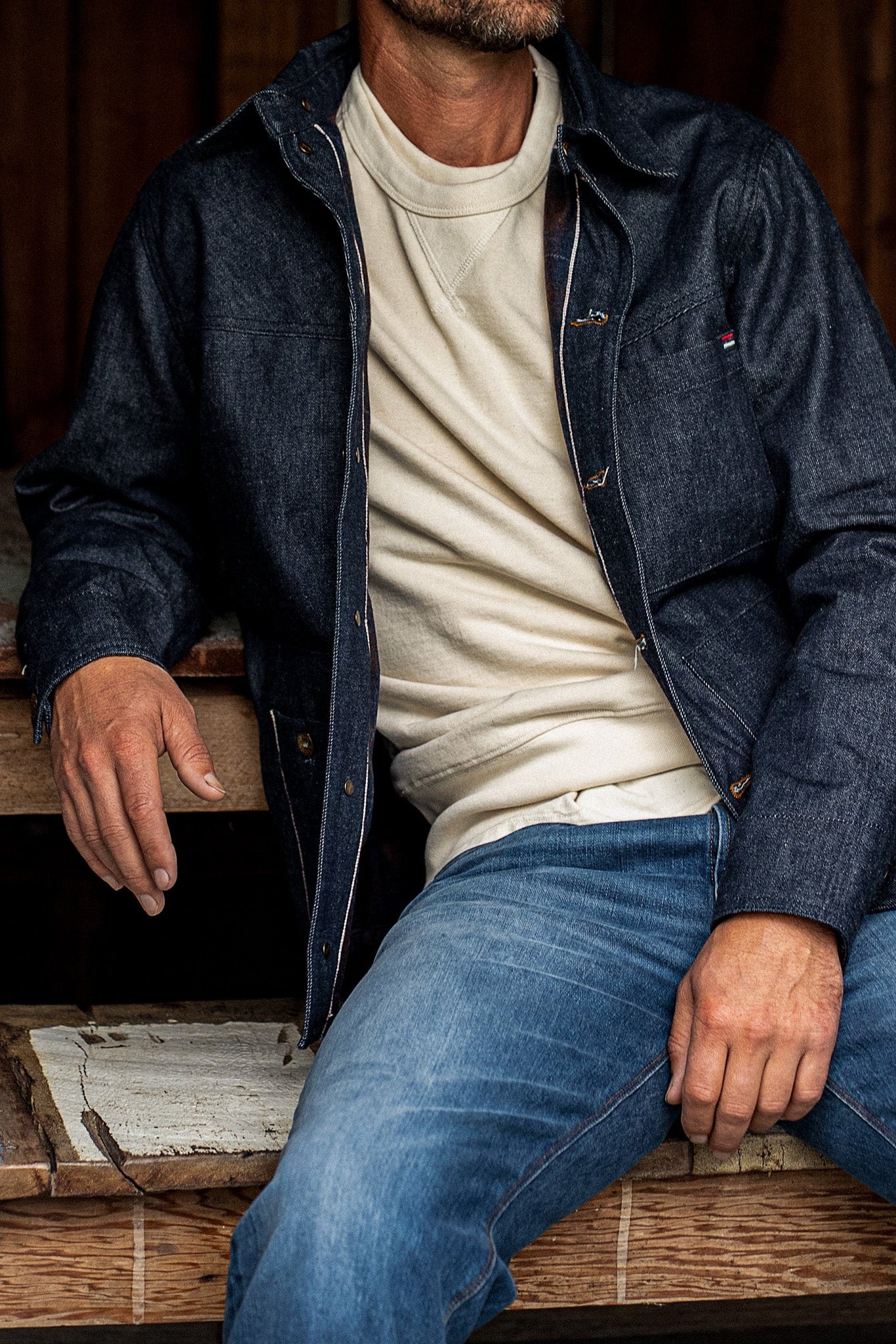 Man seated wearing the WiesMade Nico indigo denim jacket over a cream turtleneck with faded jeans.