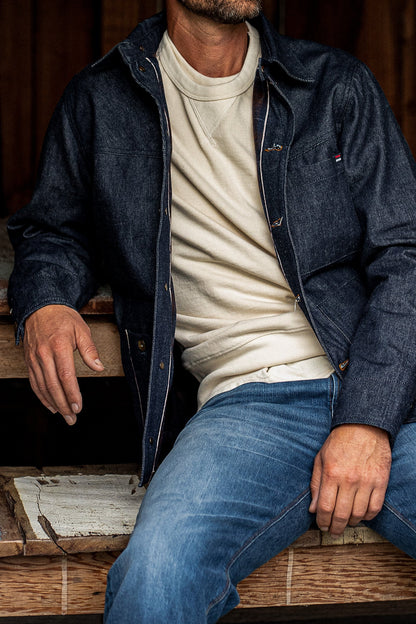 Man seated wearing the WiesMade Nico indigo denim jacket over a cream turtleneck with faded jeans.