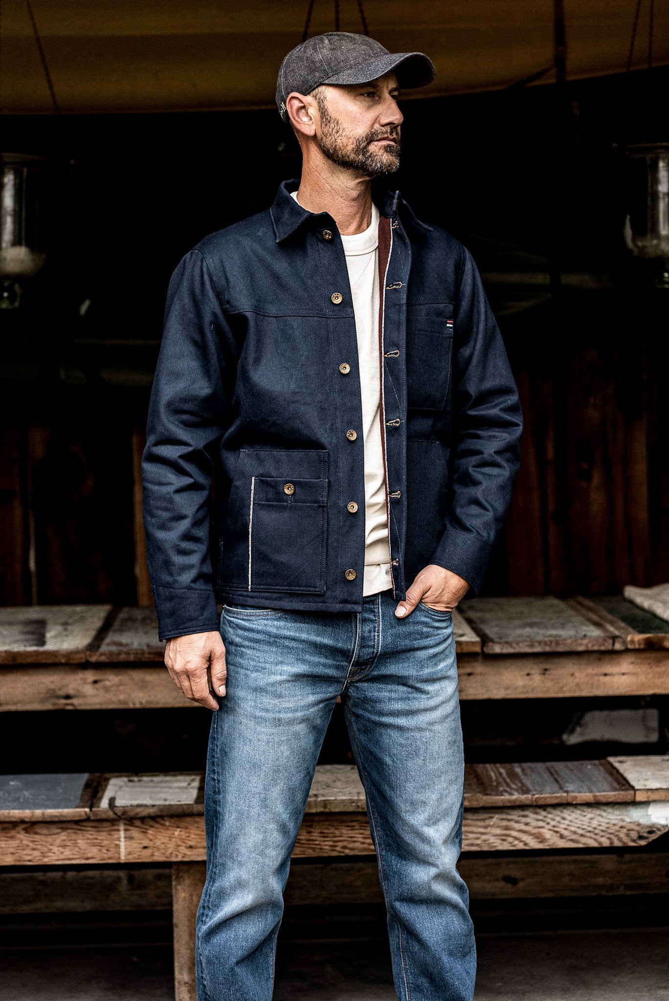 Man wearing a WiesMade Nico navy denim jacket open over a white tee with faded jeans in a rustic setting.