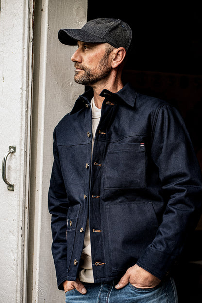 Man in a WiesMade Nico navy denim jacket and ball cap leaning in a doorway, side profile.