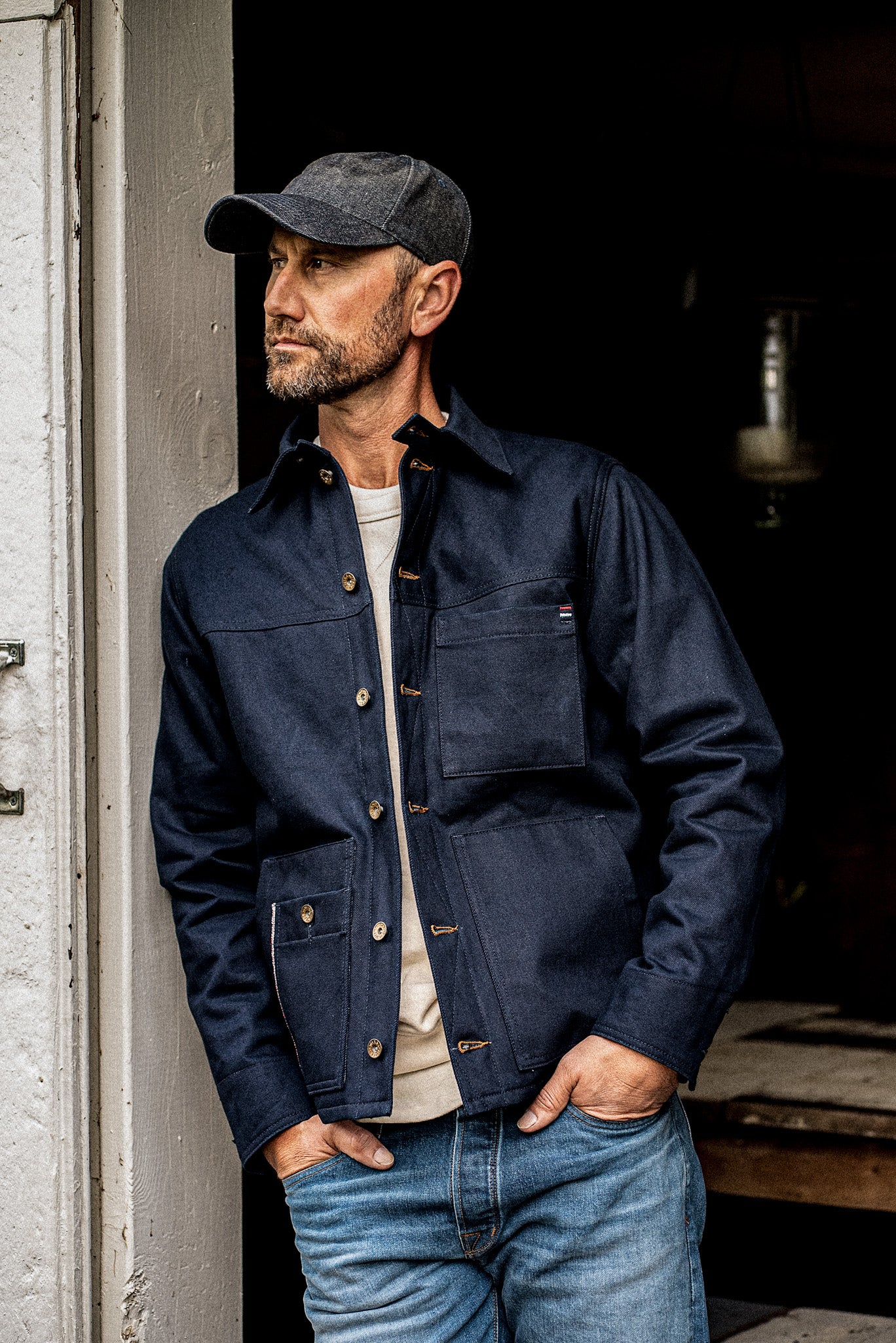 Man wearing the WiesMade Nico navy denim jacket and ball cap standing in a doorway, three-quarter view.