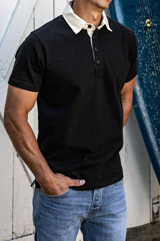 Man wearing a black short-sleeve rugby jersey with a cream collar and blue jeans beside a surfboard.
