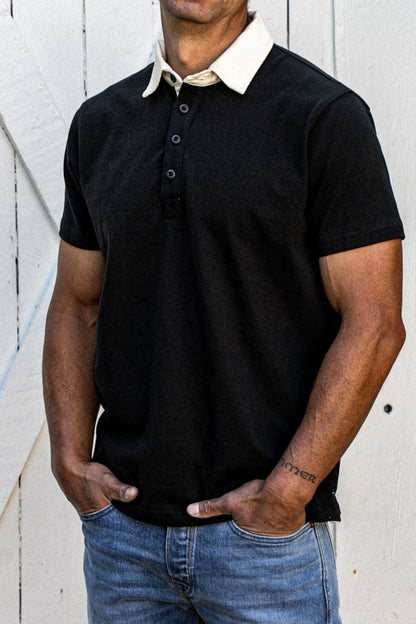 Short-sleeve black cotton rugby jersey with white contrast collar, worn with denim jeans against a white wood wall.