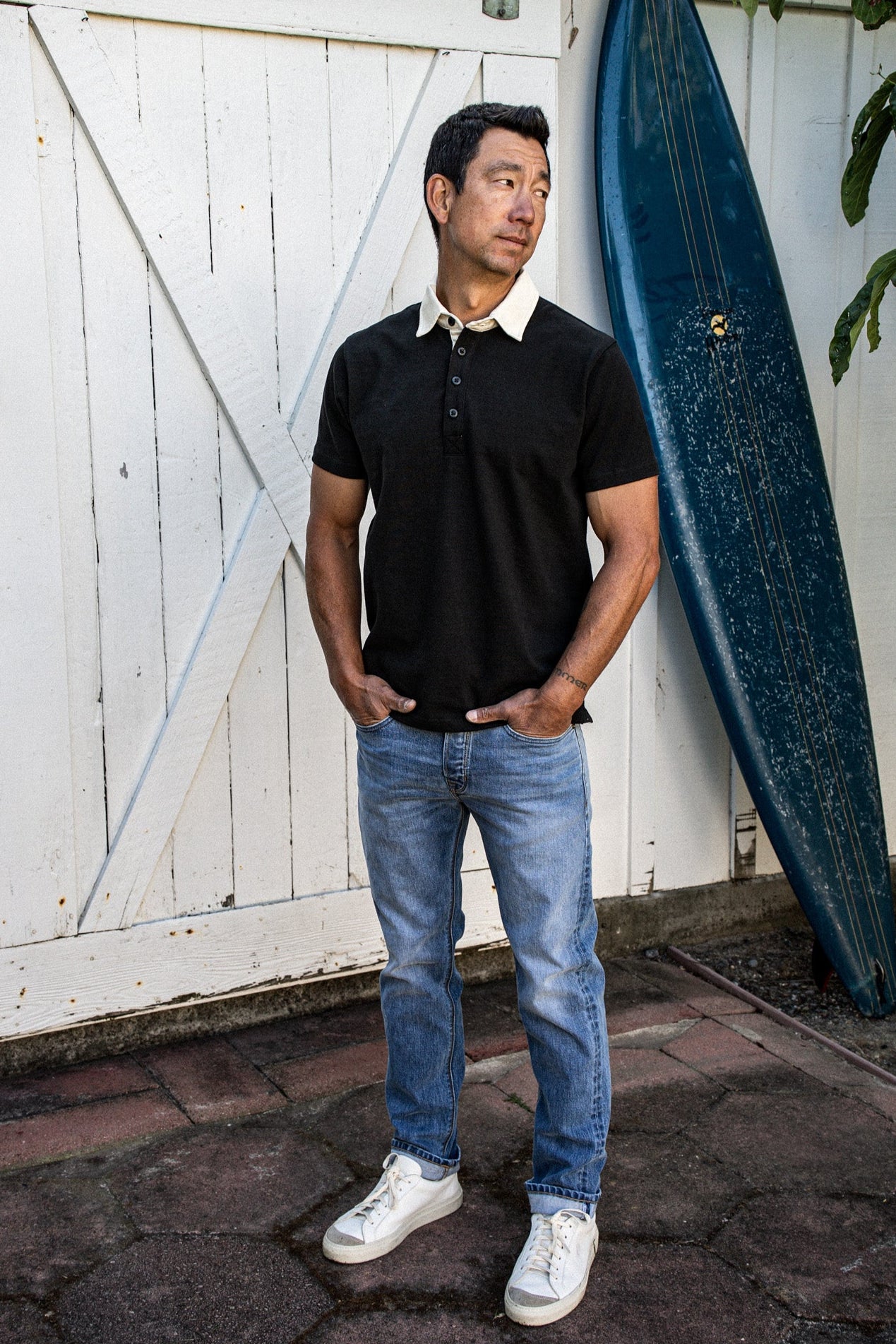 Man in a black short-sleeve rugby jersey with white collar, paired with blue jeans, standing by a barn door and surfboard.