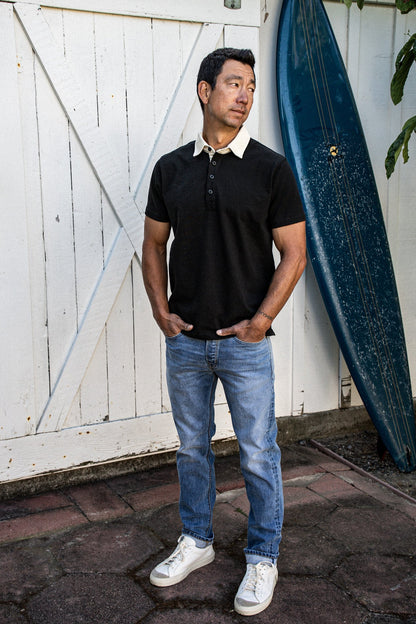 Man in a black short-sleeve rugby jersey with white collar, paired with blue jeans, standing by a barn door and surfboard.