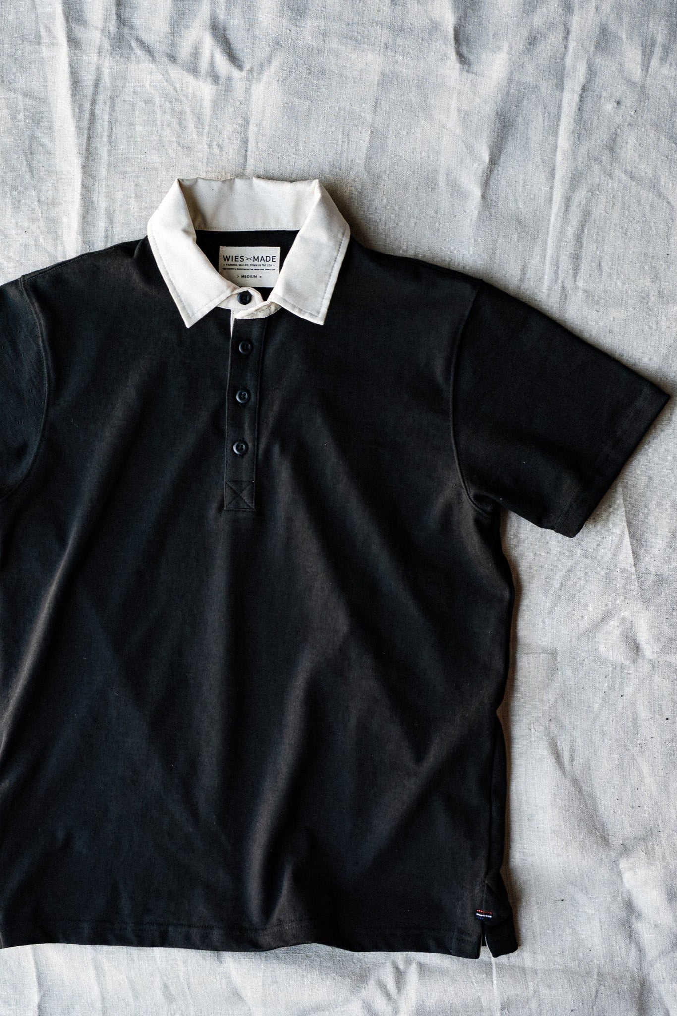 Short-sleeve basalt black rugby jersey with white collar and button placket, laid flat on linen.